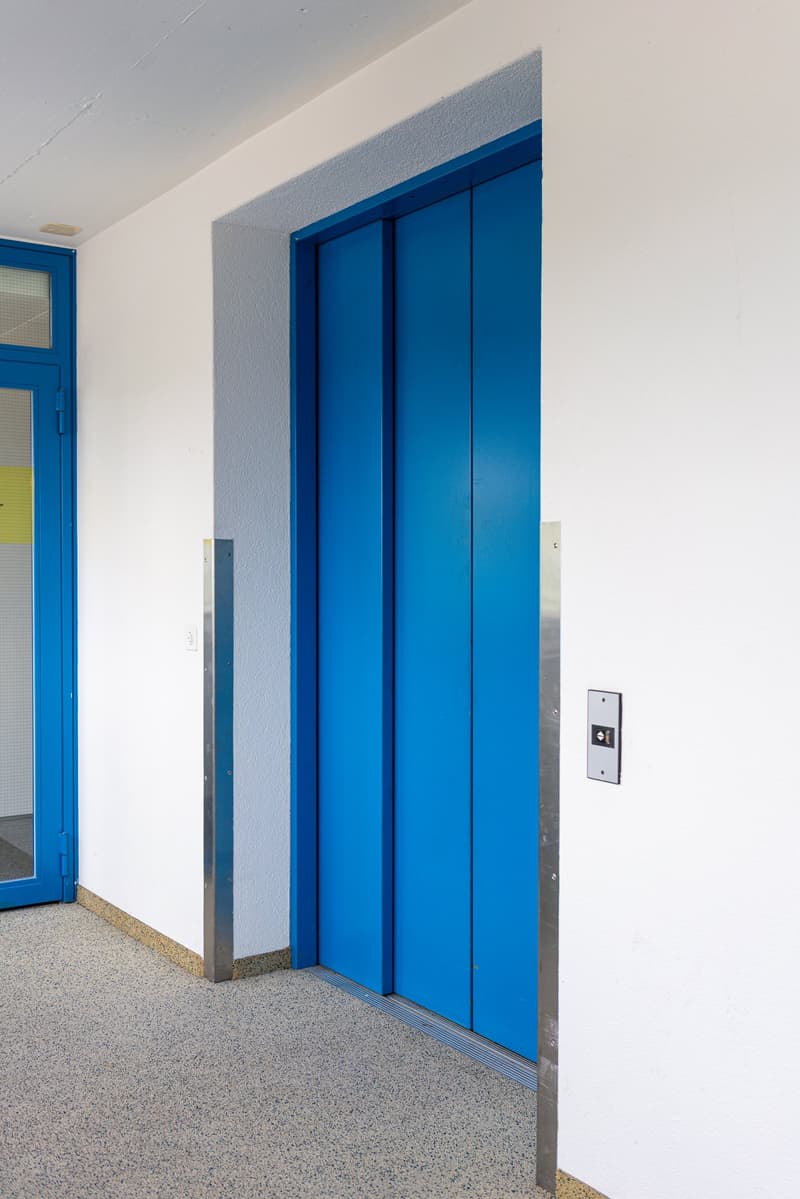 The image shows a hallway with a blue elevator door, white walls, and a terrazzo floor.