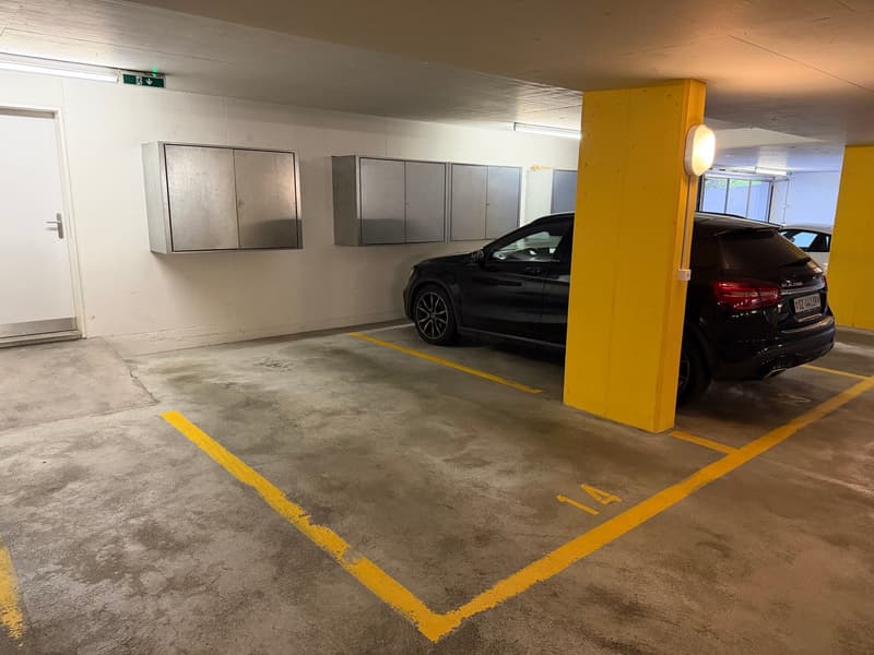 An underground parking space with yellow markings, a black car parked, and a yellow column with a light. There are two sets of metal cabinets on the wall.
