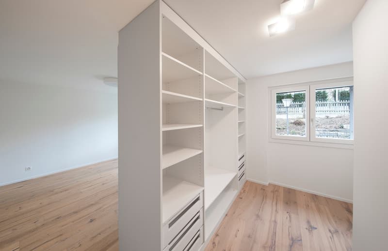 Empty room with white built-in wardrobe, wooden floor, white walls, and a window.