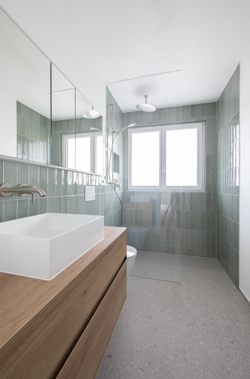 modern bathroom with white sink, wooden cabinet, green tiles, glass shower enclosure, shower head, faucet, mirror, toilet