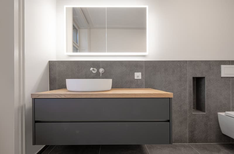 modern bathroom with white sink, wooden countertop, gray cabinet, gray tiles, mirror with light