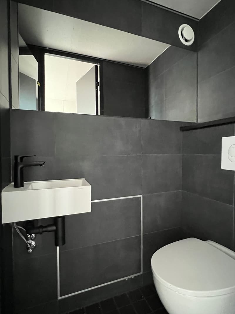 A small bathroom with dark-colored walls and floors, white sink, black faucet, white toilet, mirror, and a small shelf.