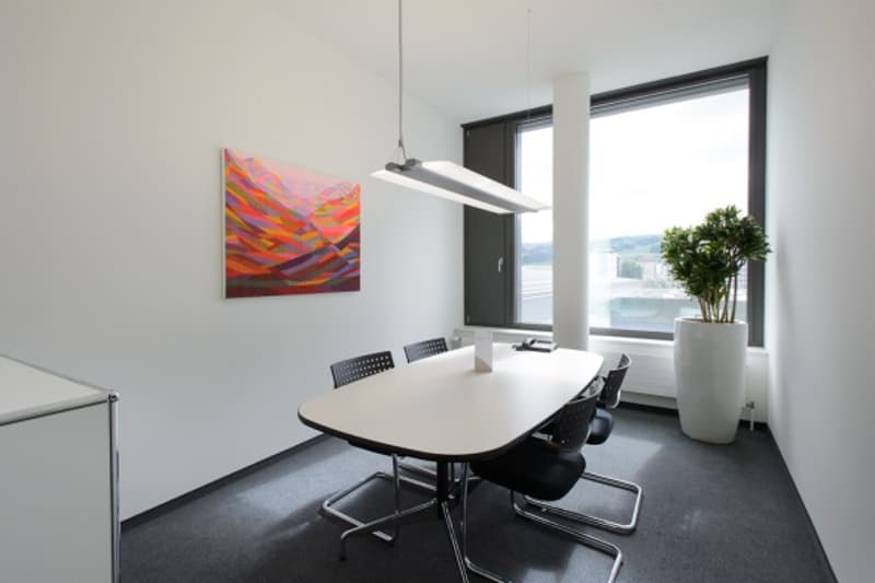 Modern office space with large windows, a rectangular conference table, black chairs, and a colorful abstract painting on the wall.