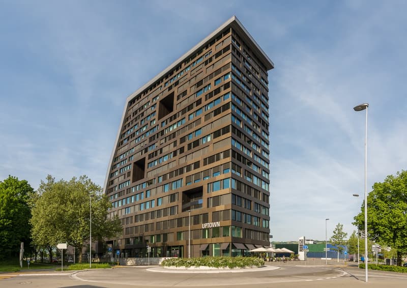 A tall, modern office building with a distinctive angular design. The building has a brown and glass facade with numerous windows. There are trees and landscaping around the building, and a street with a traffic circle in front of the building.