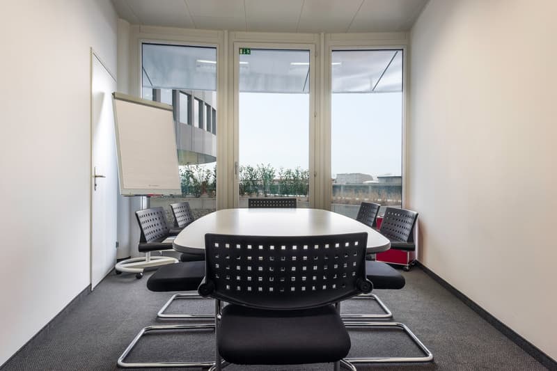 Spacious conference room with large windows providing a view, a white board, a round table with black chairs, and a carpeted floor.