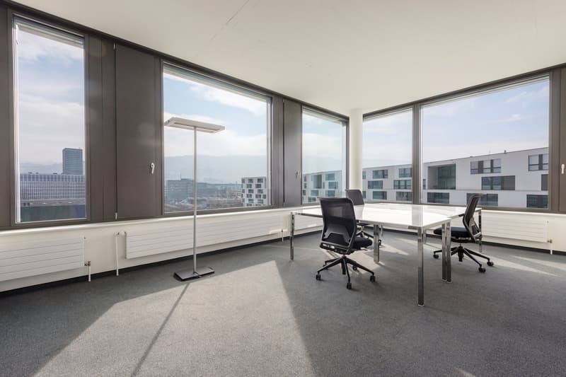 Large office space with floor-to-ceiling windows offering a panoramic view of the city skyline. The room features a long table with two office chairs, and the floor is covered in gray carpeting. The overall design appears modern and minimalist.