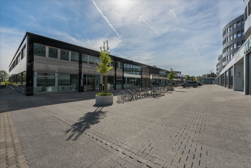 Modern office building with large windows, bicycles parked in front, paved parking area, green trees