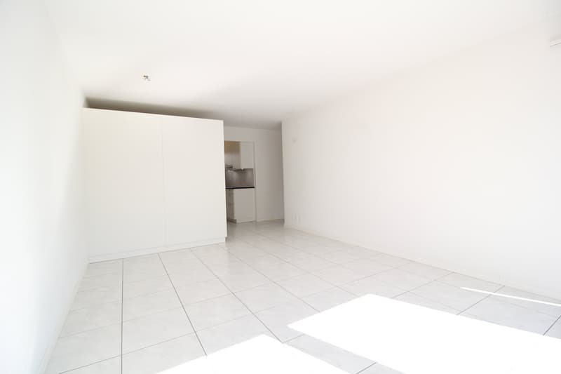 Empty room with white walls and floor, a large white cabinet, and a small kitchen area.