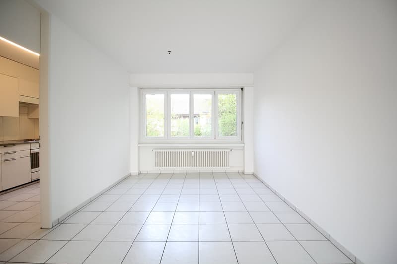 Empty room with white tiles, white walls, large window, white kitchen visible in the background
