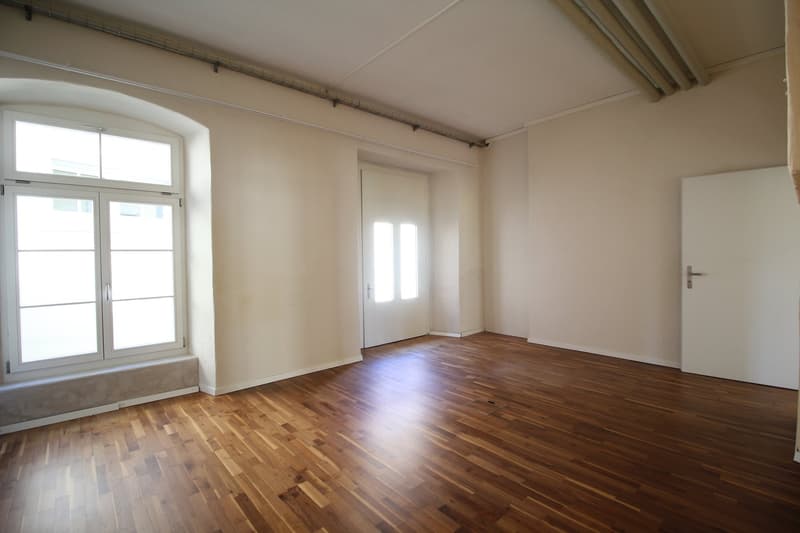 Empty room with hardwood floors, white walls, arched window, and two doors.