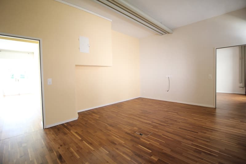 An empty room with wooden floors and beige walls. There are two doors, one open and one closed, and a power outlet on the wall. The room has a ceiling with a pipe and a square hole in the floor.