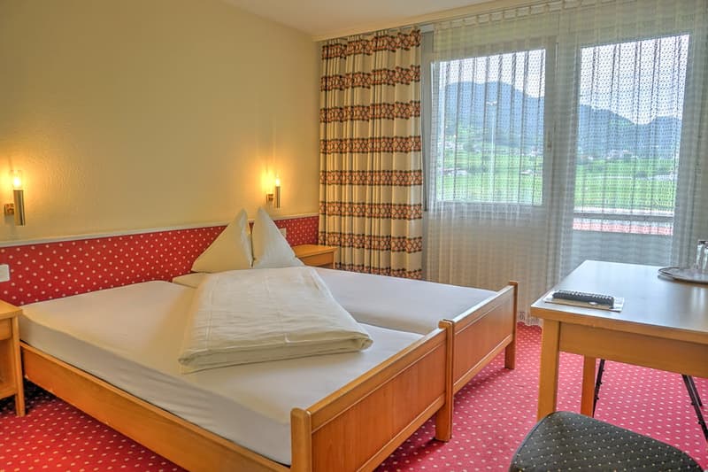 Spacious bedroom with wooden bed frame, white bedding, patterned curtains, and a desk with a TV. Large windows provide a view of the outdoor scenery.