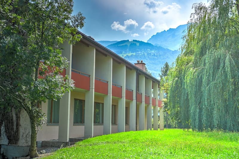 Multi-story building with colorful balconies, surrounded by lush greenery and trees, with a scenic mountain backdrop
