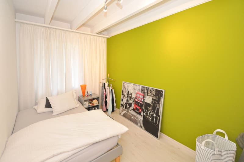 A bedroom with a single bed, white bedding, a nightstand with a lamp, and a green wall with a large picture frame.