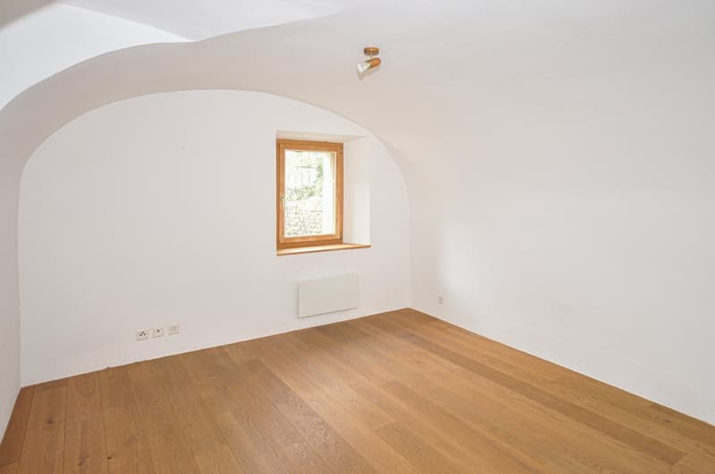 This image shows a spacious room with white walls and a wooden floor. The room has a large window that provides natural light, and there is a wall-mounted light fixture. The room appears to be empty, with no furniture or other objects visible.