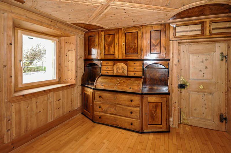 This image shows a cozy, rustic interior with wooden walls, ceiling, and floors. The room features a large, ornate wooden cabinet or dresser with drawers and cabinets. The cabinet has intricate wood carvings and detailing. There is a window that provides