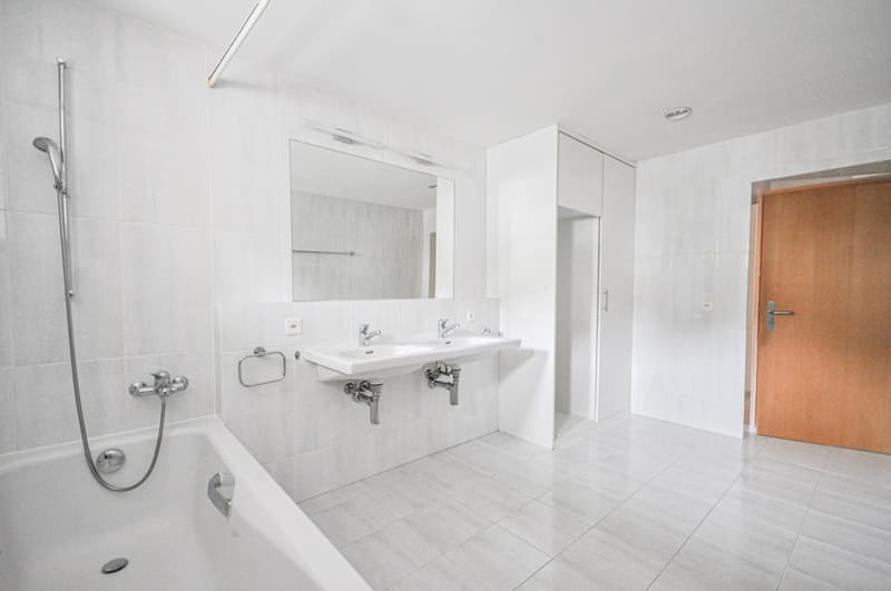 Large bathroom with white tiles, double sink vanity, mirror, and shower area