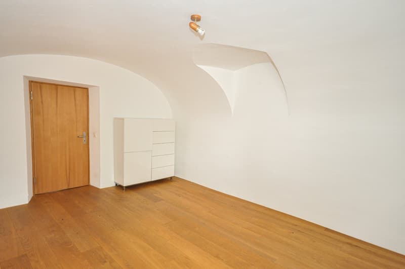 This image shows a spacious room with a wooden floor and white walls. There is a wooden door on the left side of the room, and a built-in cabinet or dresser on the right side. The room has a curved ceiling with a single spotlight fixture.