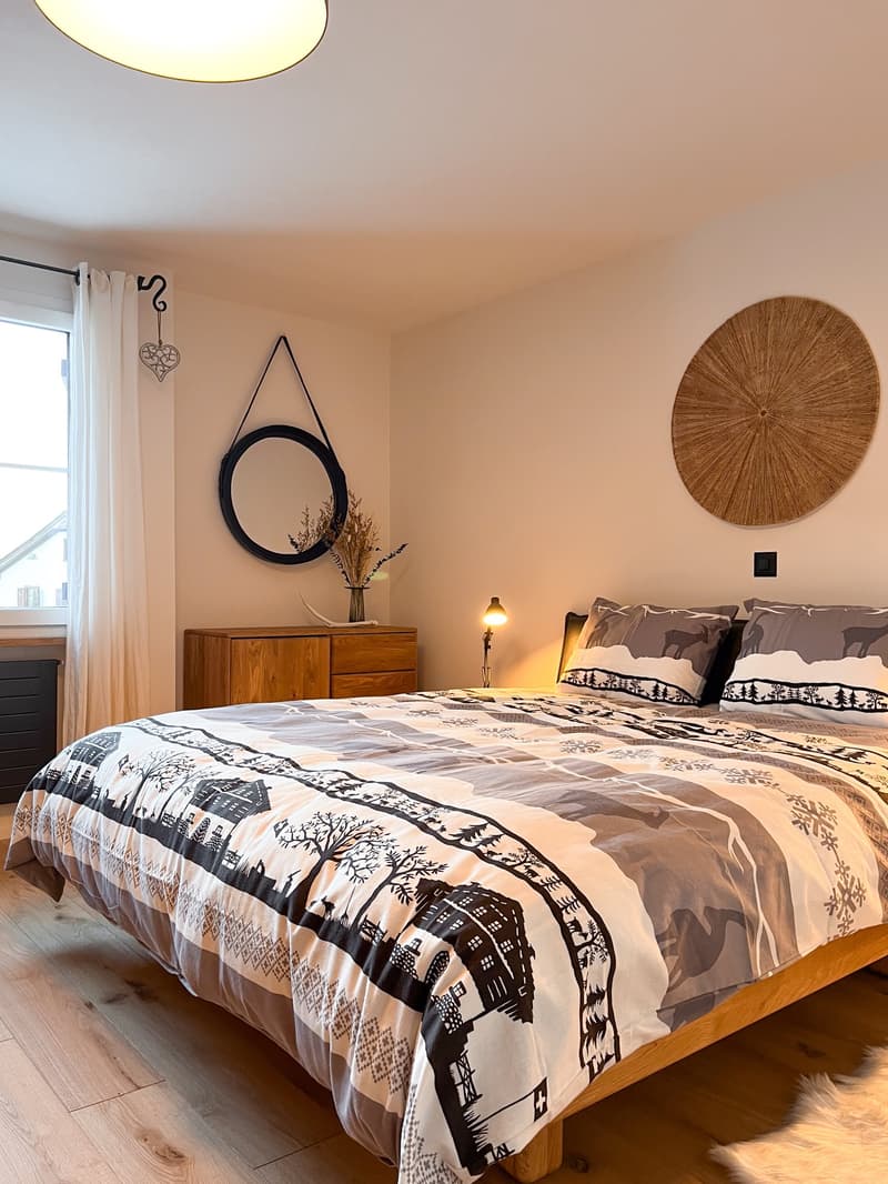 Double bed with decorative covers, bedside lamp, wooden dresser, circular mirror, decorative items, curtained window