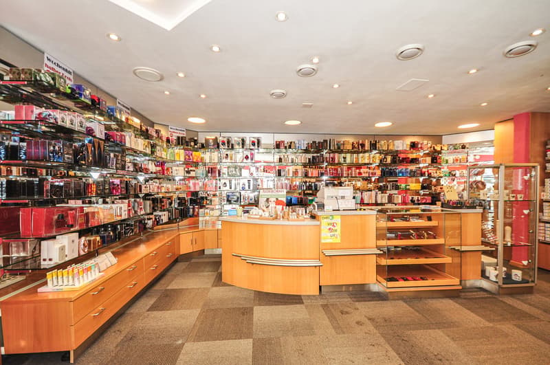 Cosmetic store with numerous shelves and cabinets full of products, a wooden reception counter with a glass top, modern lighting fixtures on the ceiling, and a checkered floor.