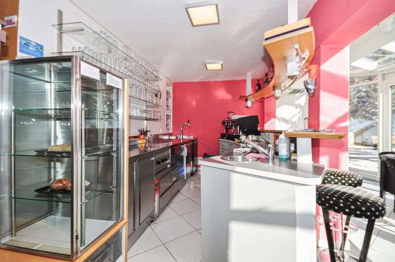 a stainless steel commercial kitchen with a glass display case, two barstools, a kitchen island, a red wall, pink lighting