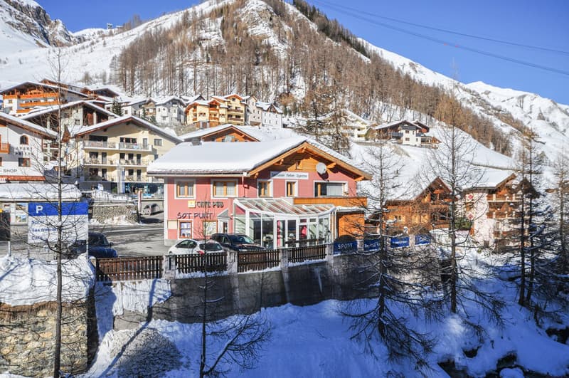 Snowy mountain area with buildings, a car park, a small stream, and mountains in the background.