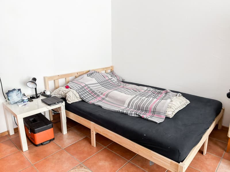 Simple bedroom with a bed, a side table, a lamp, a blanket, a pillow, and a storage box.