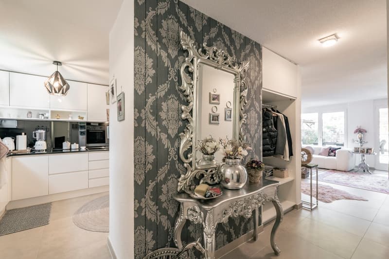 Spacious open-concept living area with a modern kitchen, ornate mirror and console table, patterned wallpaper, and a decorative lighting fixture