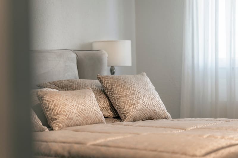 Cozy bedroom with a large bed, patterned throw pillows, and a bedside lamp. The room has a neutral color scheme and appears to have a window or balcony access.