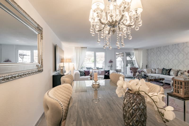 Spacious living room with large windows, hardwood floors, a grand crystal chandelier, plush beige sofas and chairs, decorative pillows, and a patterned area rug. The room has a luxurious and elegant feel.