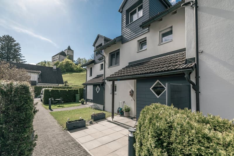 2-story house with gray siding, green roof, and large windows. There is a paved walkway leading to the entrance, with potted plants and a small garden area. A castle or tower can be seen in the background, suggesting this is a residential property near a