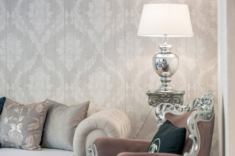 The image shows a living room with a patterned wallpaper, a sofa with decorative pillows, and a silver lamp on an ornate side table.