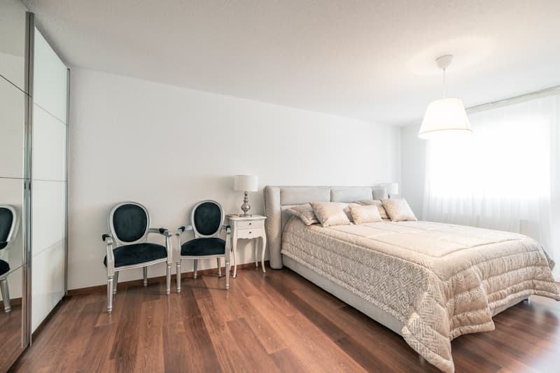 Spacious bedroom with hardwood flooring, white walls, large windows, a king-size bed with a patterned comforter, two black armchairs, and a nightstand with a lamp.