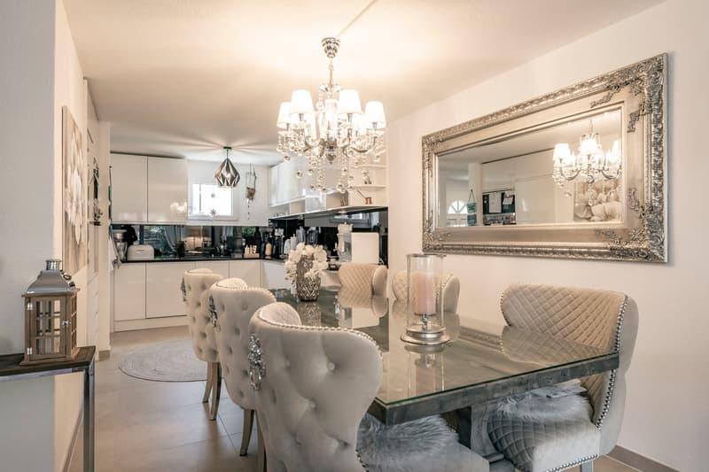 Elegant dining room with a large glass dining table, tufted upholstered chairs, a crystal chandelier, and a decorative mirror on the wall. The kitchen is visible in the background.