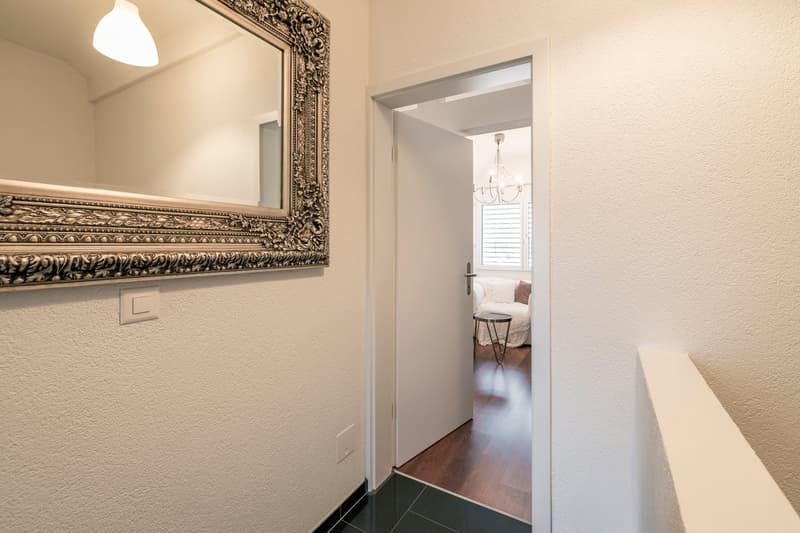 Hallway with a large ornate mirror, hardwood floors, and a doorway leading to a bedroom with a chandelier visible.