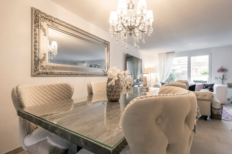Elegant dining room with a large ornate mirror, a glass-topped dining table with upholstered chairs, and a crystal chandelier hanging from the ceiling.