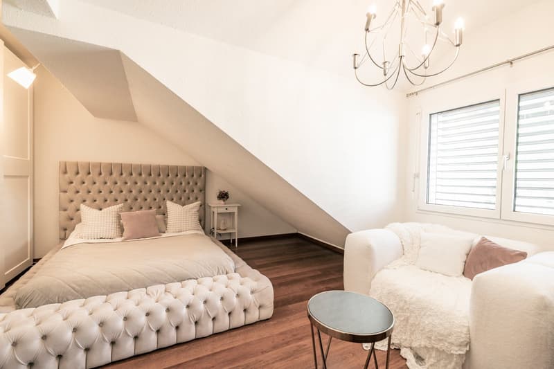 Spacious bedroom with a tufted upholstered headboard, hardwood flooring, a chandelier, and a cozy seating area by the window.