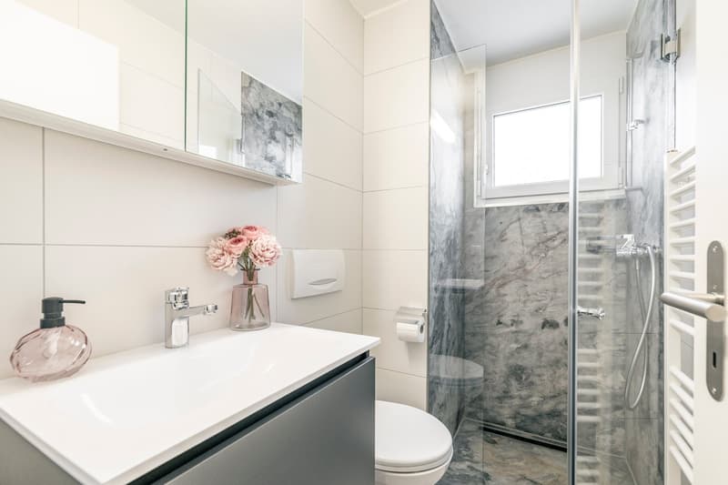 Modern bathroom with white tiles, marble-like wall panels, a vanity with a white countertop, a toilet, and a glass-enclosed shower area.