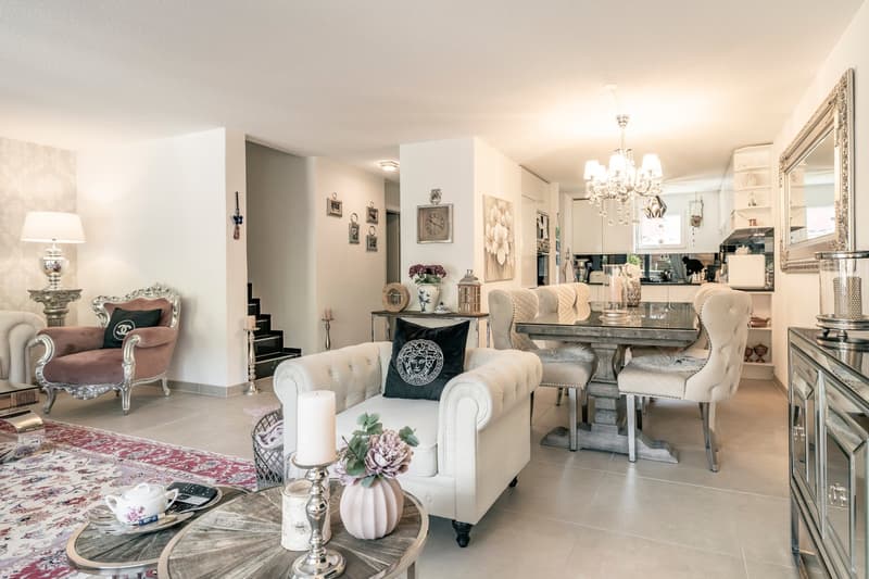 Spacious open-concept living and dining area with elegant decor. The room features a large chandelier, tufted white sofas, a mirrored coffee table, and a dining table with upholstered chairs. The walls are decorated with framed artwork and shelves. A fire