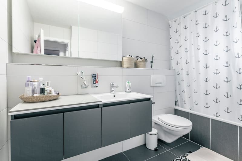 Modern bathroom with white and gray tiles, a vanity with a sink and storage cabinets, a toilet, and a patterned curtain with anchor designs on the wall.