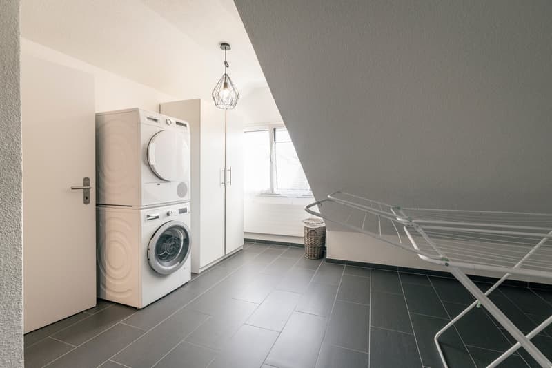 Spacious laundry room with a washing machine and dryer, a drying rack, and a pendant light fixture. The room has gray tile flooring and white walls.