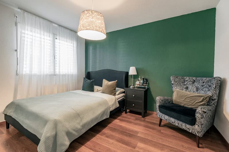 Spacious bedroom with green accent wall, large windows, hardwood flooring, black upholstered bed frame, nightstand, and patterned armchair