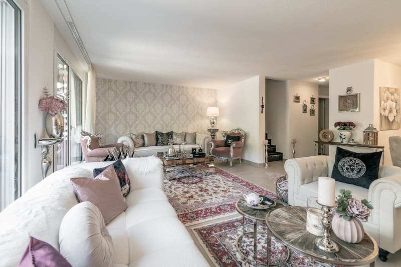 Spacious living room with large windows, patterned wallpaper, plush sofas and armchairs, ornate coffee table, decorative accents like vases, candles, and artwork on the walls