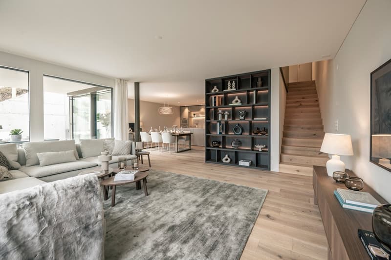 Spacious open-concept living area with large windows, hardwood floors, grey sofa, coffee table, and a built-in shelving unit displaying decorative items. The room also features a dining area and kitchen visible in the background.