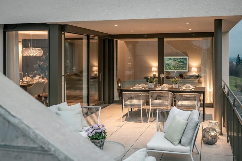 Spacious open-plan living and dining area with large sliding glass doors leading to a balcony or terrace. The room features a comfortable sofa, a dining table with rattan chairs, and a decorative chandelier. The space is well-lit and has a cozy, modern ae