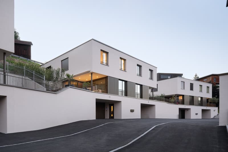 3-story modern apartment building with gray exterior, large windows, balconies, and a parking area in the front