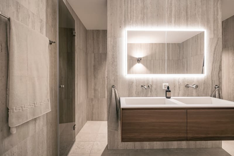 Large bathroom with marble-like tile walls, double vanity with wooden cabinets, large backlit mirror, and shower stall