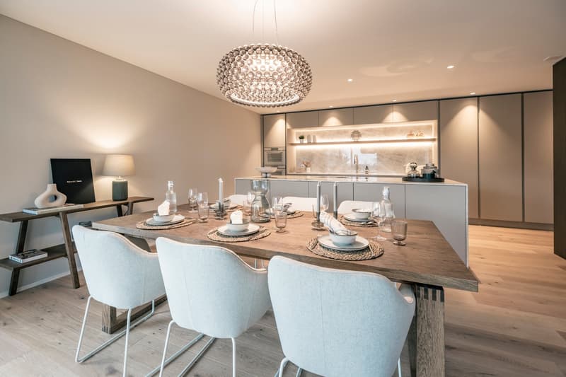 Spacious dining area with a large wooden dining table, white upholstered chairs, and a modern crystal chandelier. The kitchen is visible in the background, featuring gray cabinets and a marble backsplash. The room has a warm, contemporary design with hard