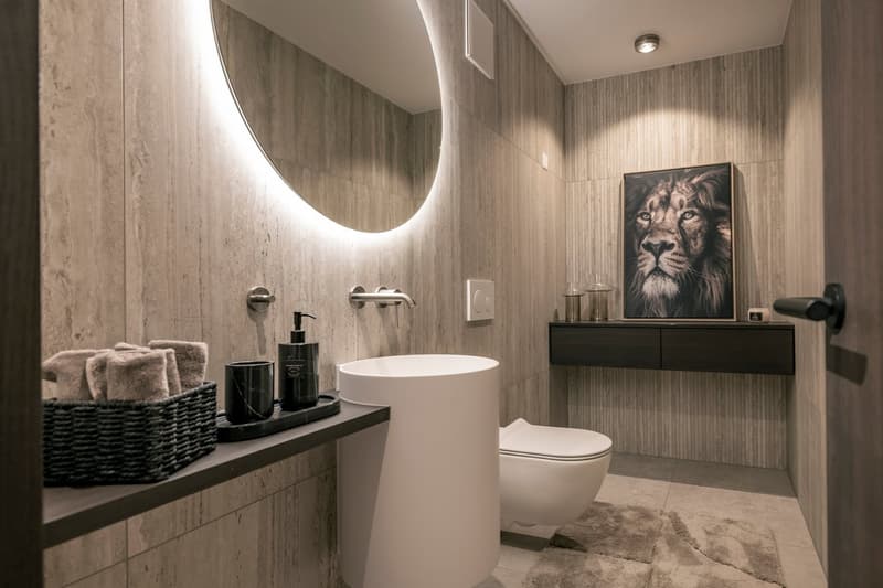 Modern bathroom with gray tiled walls, a round backlit mirror, a white freestanding bathtub, a black vanity with a vessel sink, and a large framed artwork of a lion on the wall.