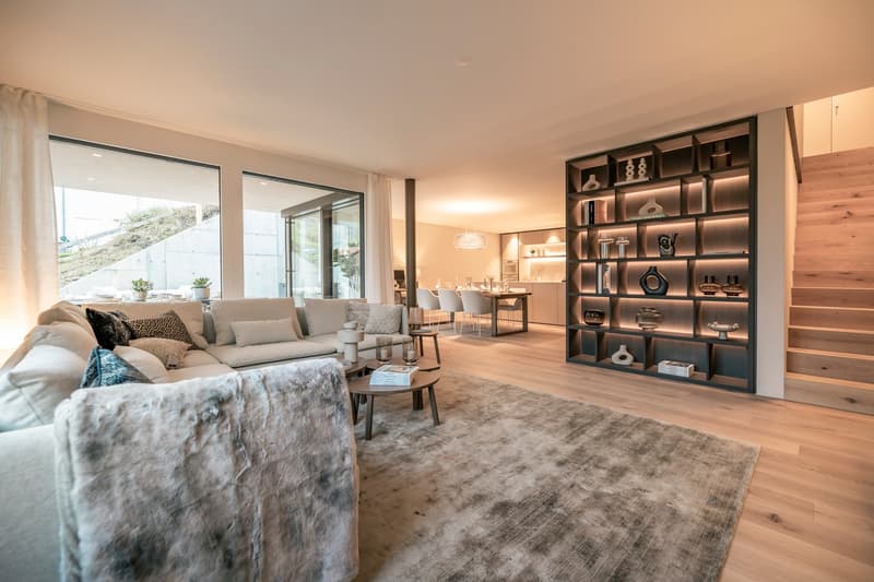 Spacious open-plan living room with large windows, plush gray sectional sofa, wooden shelving unit with decorative items, dining area with table and chairs, hardwood flooring, and a cozy rug.